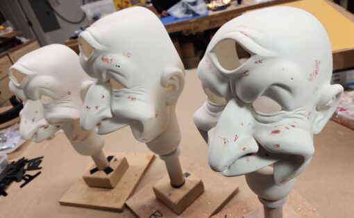 Three unfinished sculpted masks on stands.